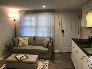 Ein Wohnzimmer mit einer Couch, einem Hocker und einem kleinen Tisch mit Tablett. Ein Fenster mit Vorhängen und eine Tür zur Rechten sind zu sehen.