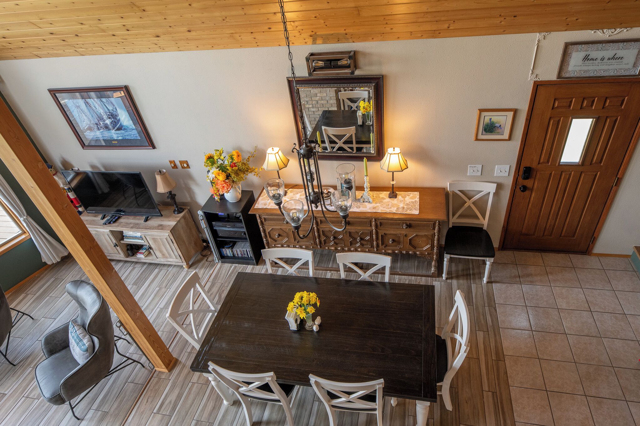 Un comedor con una mesa de madera oscura, sillas blancas y un candelabro. Hay un televisor, una mesita con un florero y un espejo en la pared.