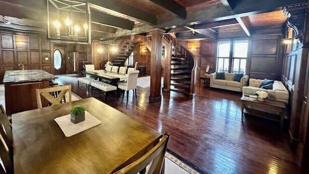A spacious living room with a wooden staircase, a dining area with a large wooden table, and a cozy seating area with a sofa and armchairs.