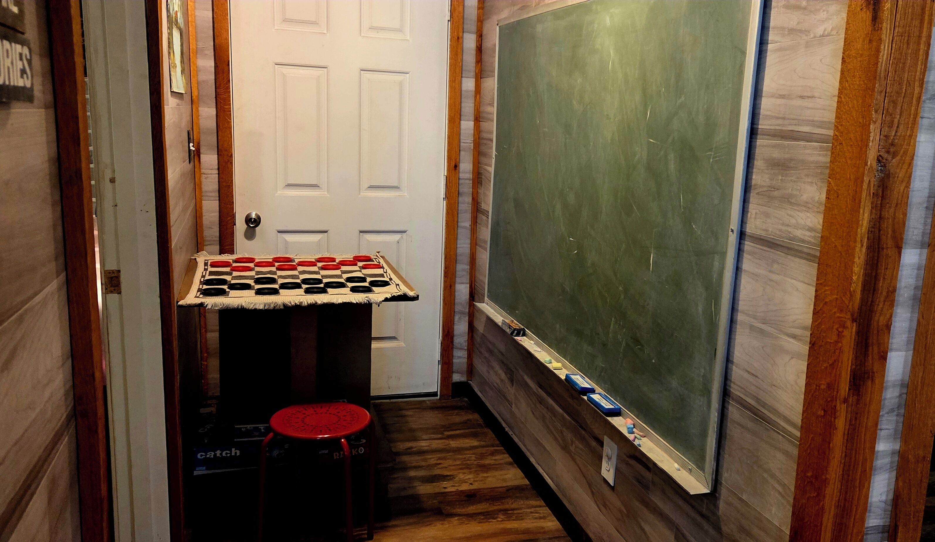 A narrow hallway with a chess table and stools, a door, and a chalkboard wall.