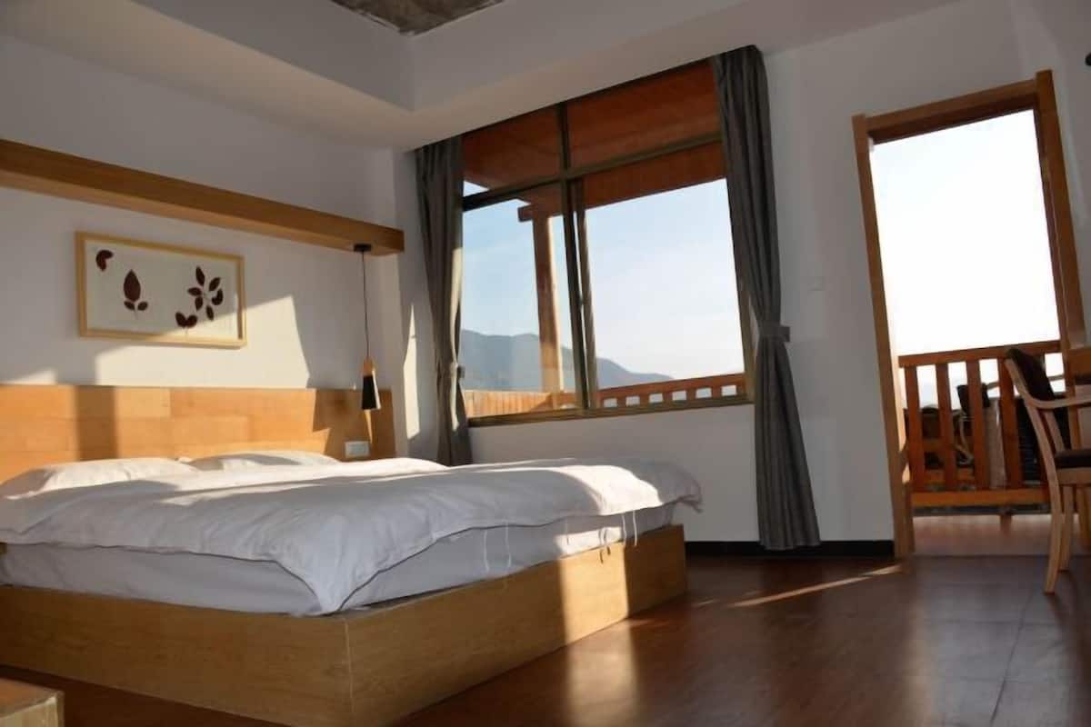 A bedroom with a wooden bed, a large window with curtains, a wooden chair, and a framed picture on the wall.