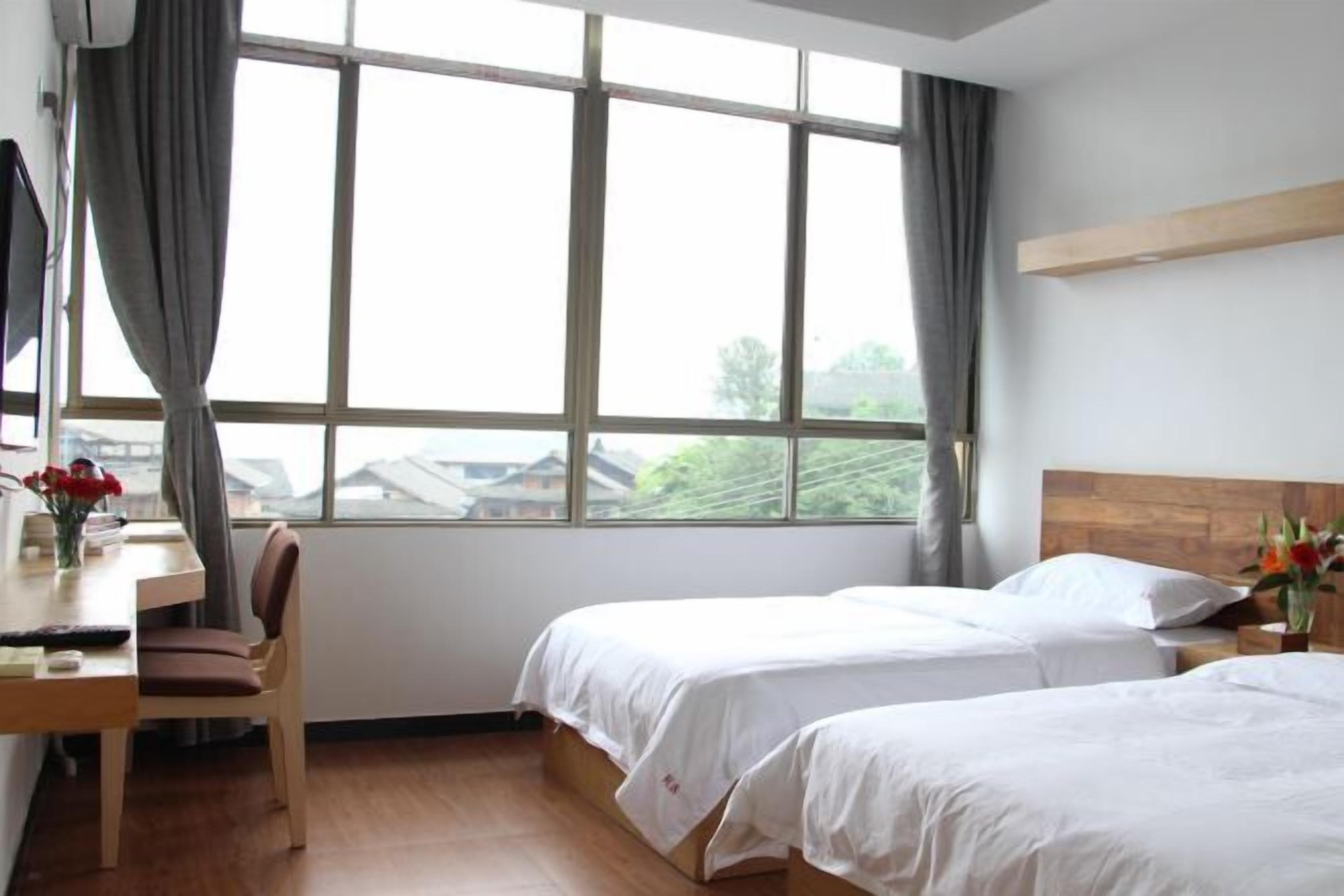 A hotel room with two beds, a desk with a chair, and a large window with curtains.