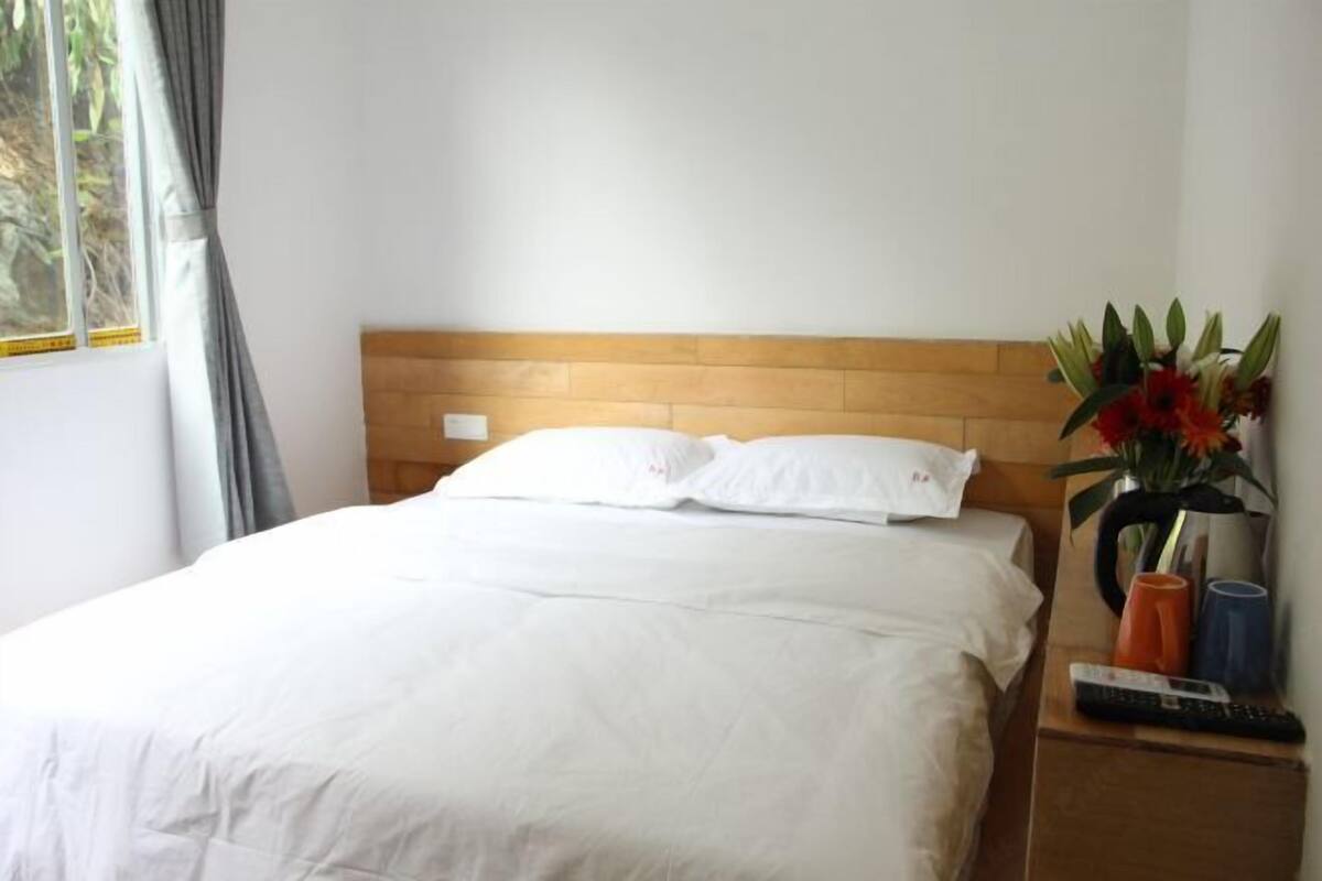 A neatly made bed with white linens, a wooden headboard, a nightstand with a remote control, a vase with flowers, and a window with curtains.