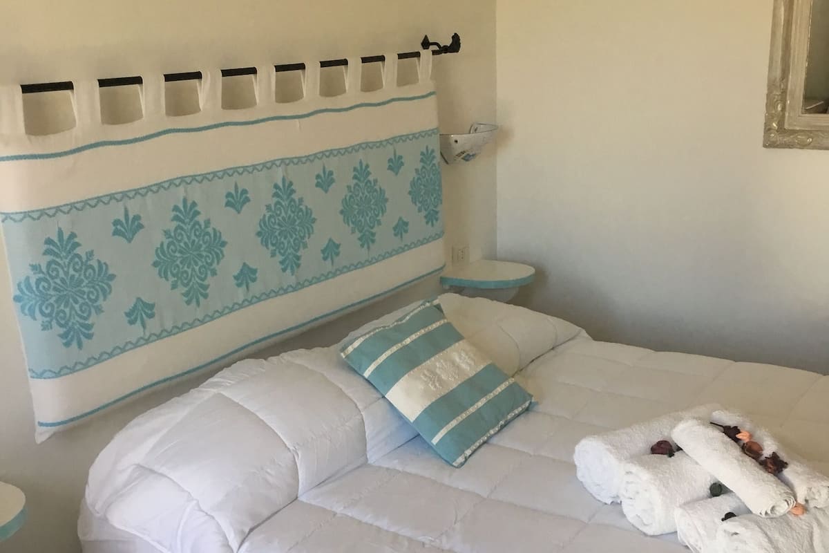 A neatly made bed with white bedding and a blue and white headboard. There are rolled towels on the bed, and a blue rug on the floor.