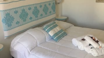 Ein ordentlich bezogenes Bett mit weißer Bettwäsche und einem blau-weißen Kopfteil. Auf dem Bett liegen zusammengerollte Handtücher, und auf dem Boden liegt ein blauer Teppich.