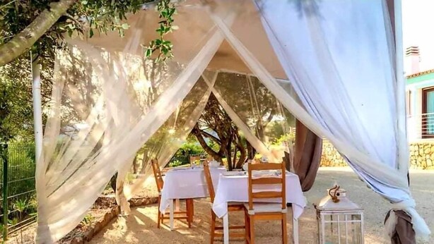 An outdoor dining area with tables and chairs under a tent-like structure, surrounded by trees. - Tenuta Petra manna  (Palau)