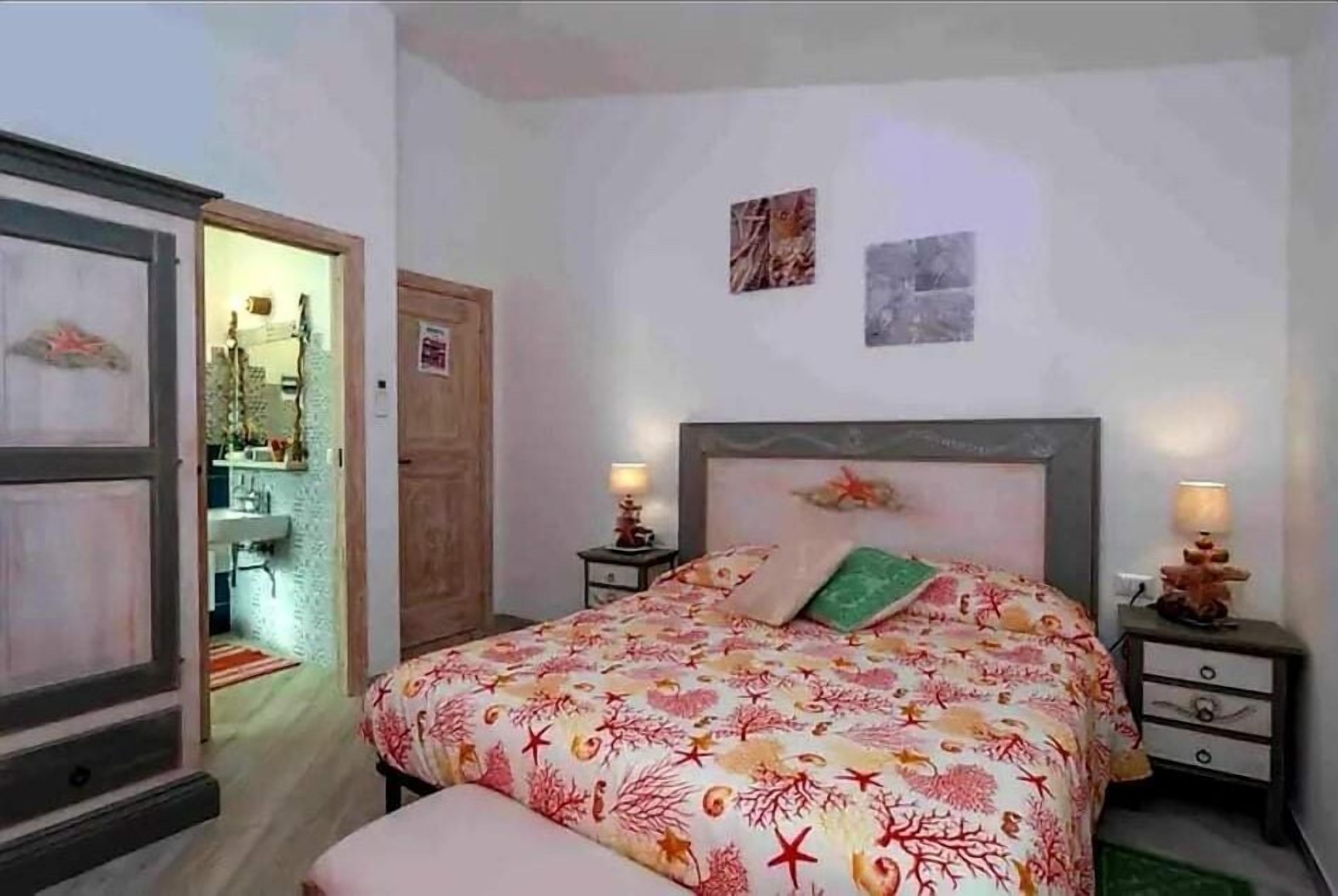 A bedroom with a bed, two bedside tables, a nightstand, a door, and a small bathroom visible through an open door.
