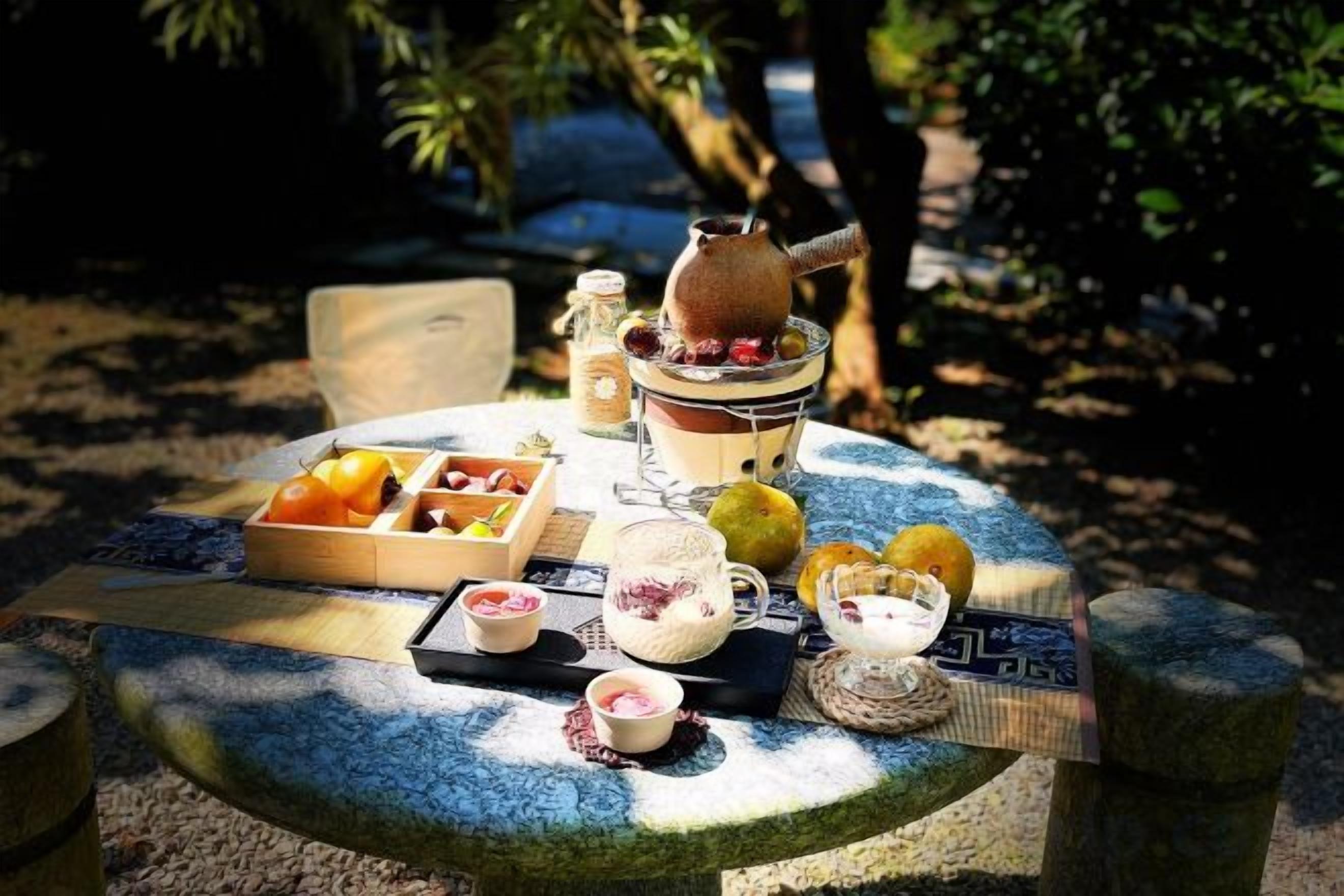A round table with a variety of food items and a teapot, set outdoors with a natural backdrop.