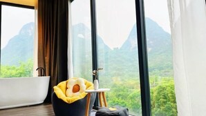 Una stanza con una vasca da bagno, un'ampia finestra vista montagna, un cuscino a forma di fiore giallo e un tavolino con un vaso.