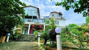 Edificio residencial de dos pisos con balcón, entrada principal con escalones y área de jardín con plantas y un sendero.