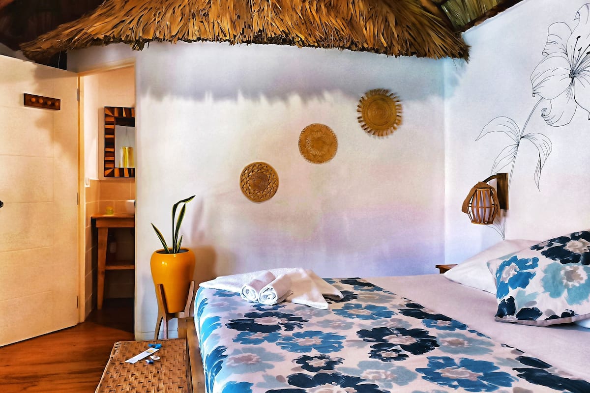 A room with a bed, a wall with decorative plates, a potted plant, and a thatched roof.