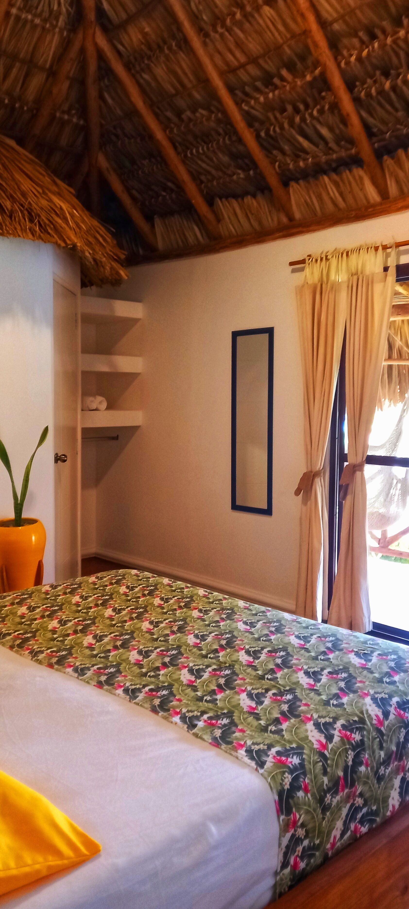 A room with a bed, a potted plant, a mirror, and a window with curtains.