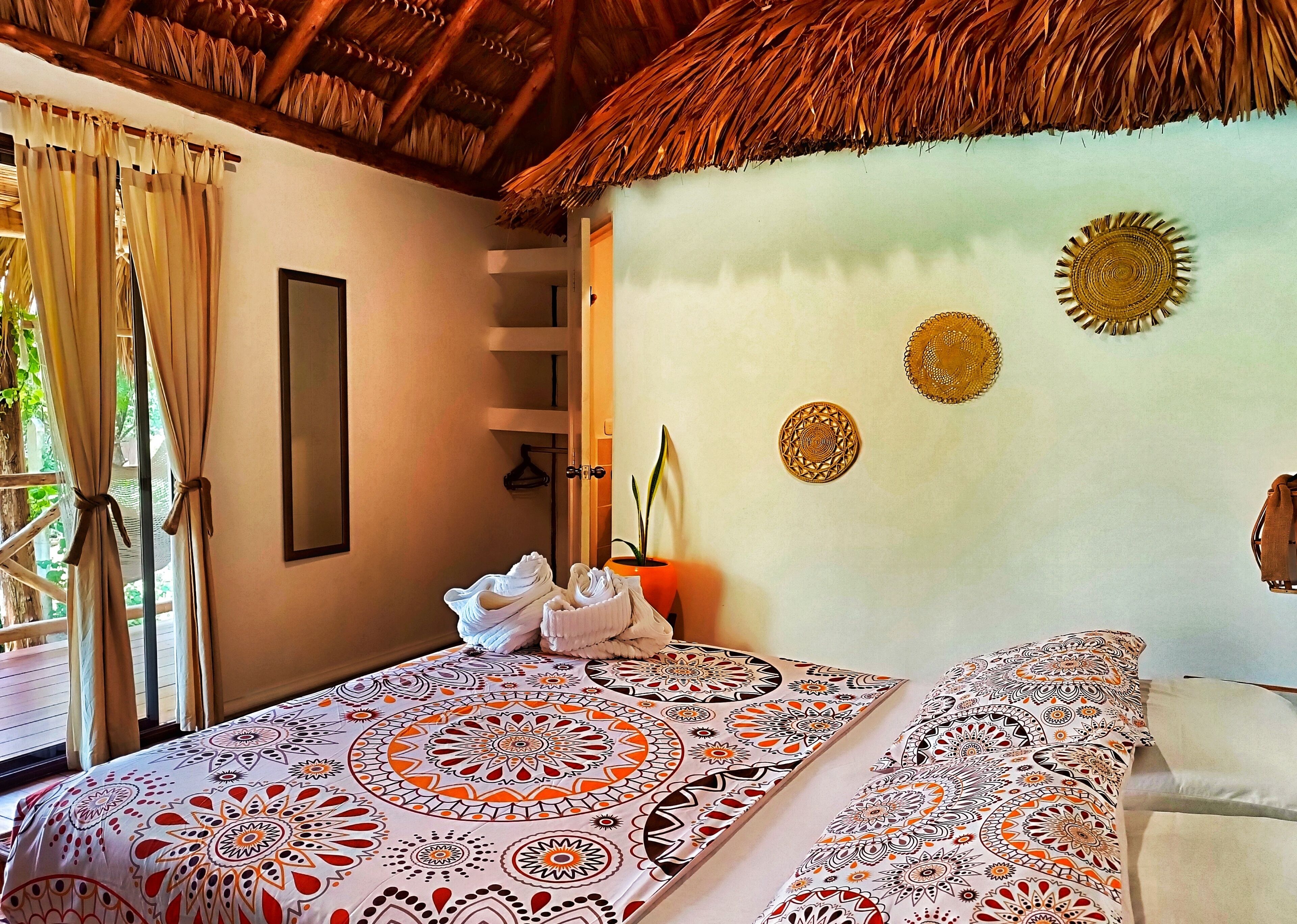 Una habitación con cama, ventana con cortinas, techo de paja y decoración en las paredes.