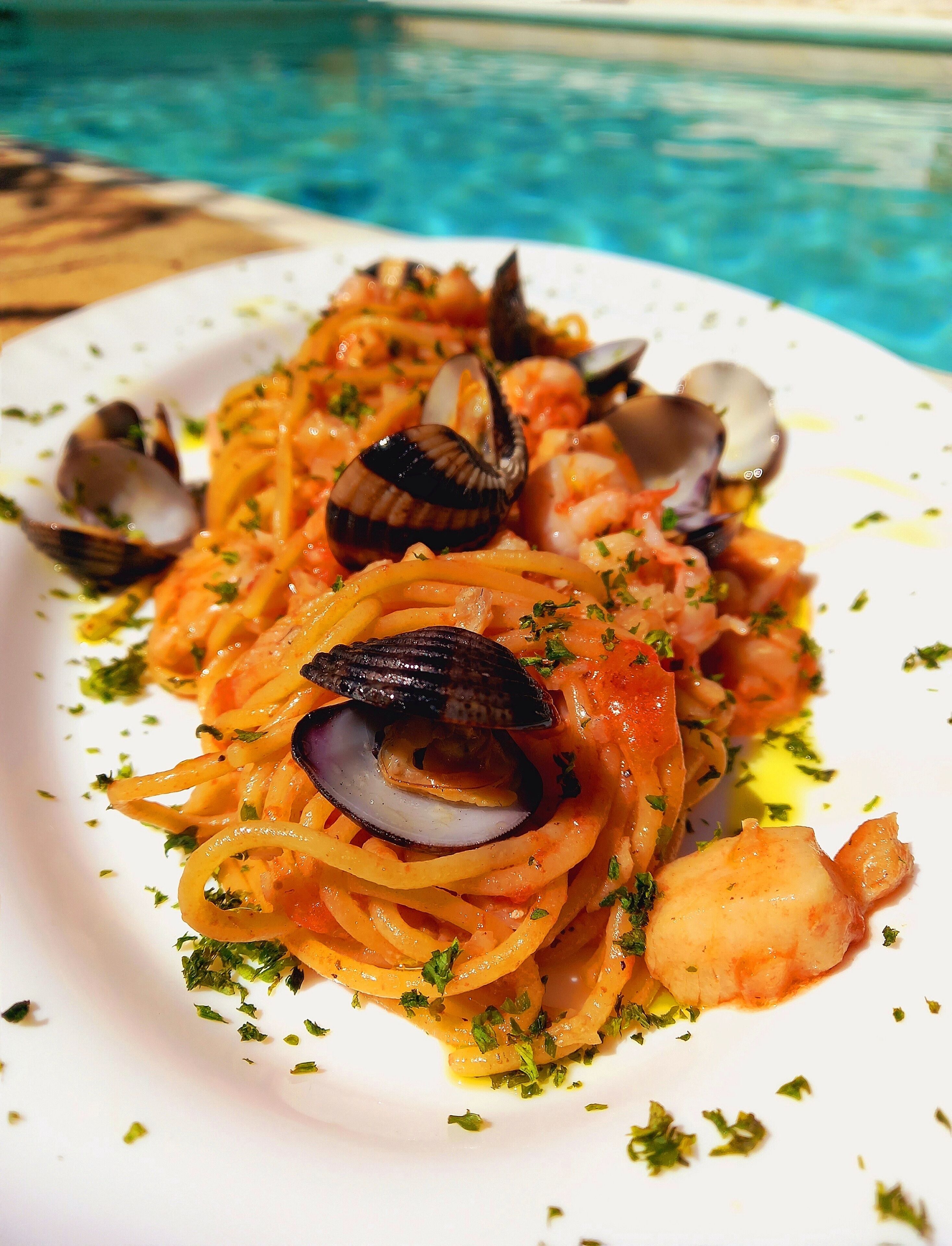 Une assiette de pâtes aux fruits de mer, avec palourdes et crevettes, garnie d’herbes aromatiques, servie au bord d’une piscine.