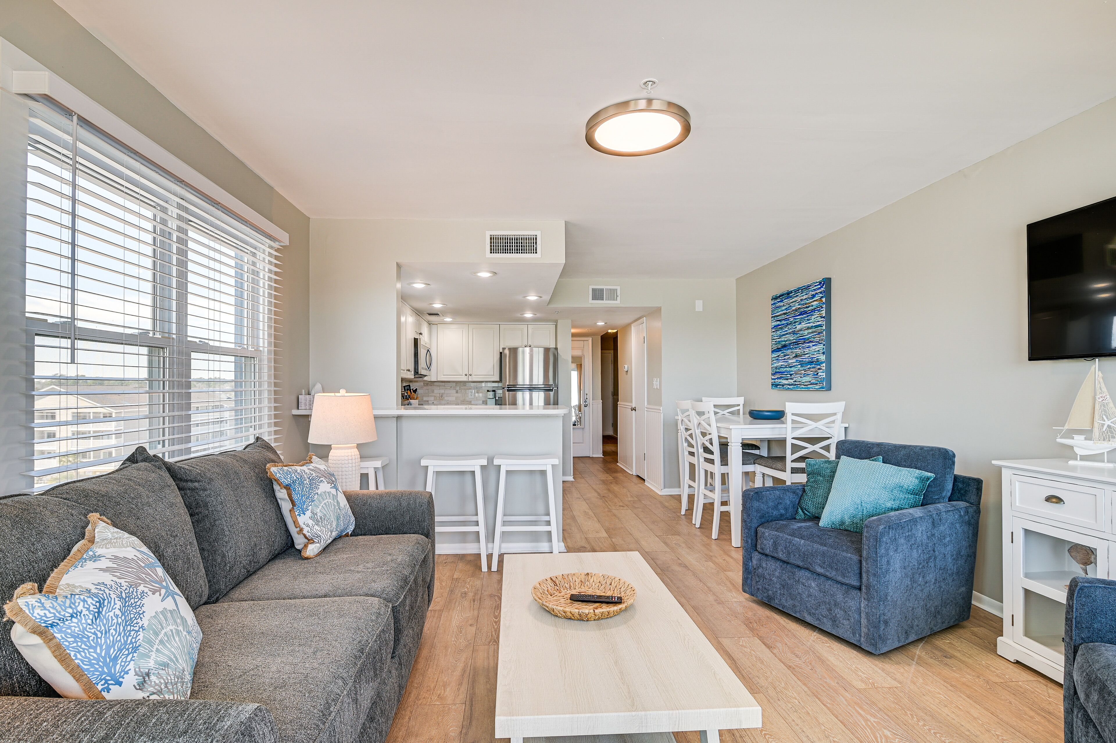 A modern living room with a grey sofa, a blue armchair, and a white coffee table. A kitchen with white cabinets and a dining area with a white table and chairs are visible in the background.