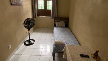 A small, single-room apartment with a bed, a table, a fan, and a window with curtains.
