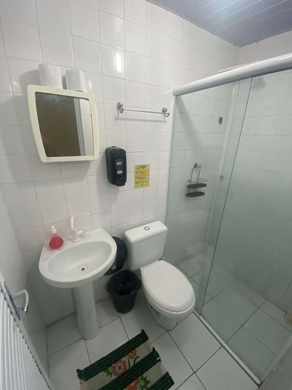 Un baño con lavamanos, inodoro y ducha blancos. Hay un espejo, dispensador de jabón y un letrero amarillo en la pared.