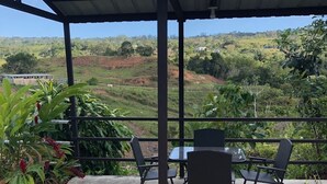 A covered outdoor seating area with a table and chairs, surrounded by greenery and a view of a hilly landscape. - comfortable, fresh, 2 bedroom in the country (Utuado)
