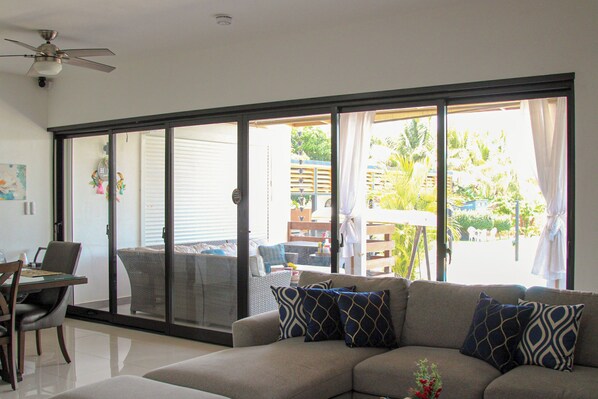 Una sala de estar con un sofá, una mesa de comedor y sillas. Hay un ventanal corredero que da a un área exterior con patio.