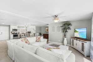 A modern living room with a white sofa set, a dining area with a kitchen in the background, a ceiling fan, and a television displaying a beach scene.