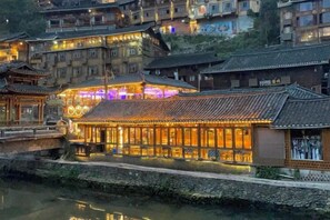 A traditional building with multiple levels, a river in the foreground, and a well-lit facade. - Xijiang Anxia B&B (Leishan)
