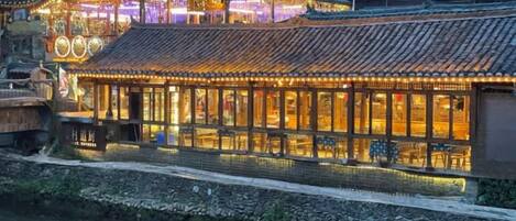 A traditional building with a tiled roof, a waterway in front, and a well-lit interior.