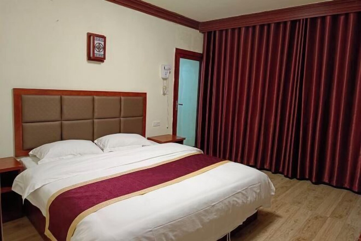 Quarto de hotel com uma cama grande, cabeceira de madeira, cortina vermelha e uma pequena mesa de cabeceira.