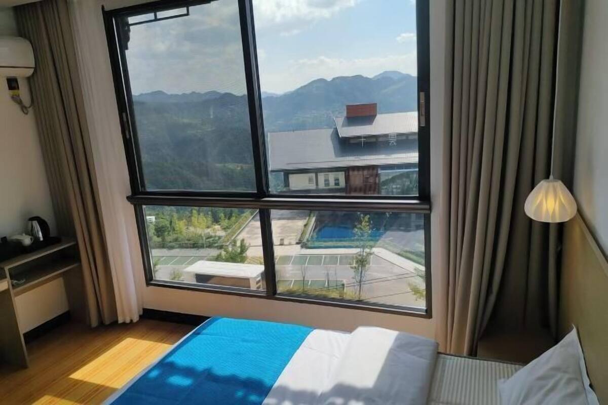 A hotel room with a large window offering a mountain view, a bed with a blue and white bedspread, a bedside table with a lamp, and a view of a pool and building in the background.