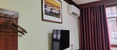 Quarto com escrivaninha de madeira, televisão fixada na parede, ar condicionado, quadro emoldurado e janela com cortinas.
