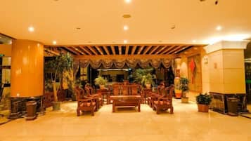 A lobby with a central wooden bench surrounded by chairs, potted plants, and a decorative ceiling.