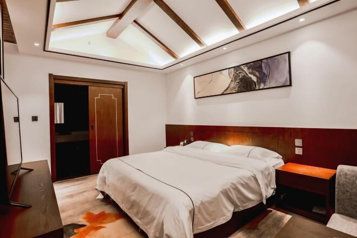 A modern bedroom with a skylight, a bed with white linens, a wooden headboard, a bedside table, and a painting on the wall.