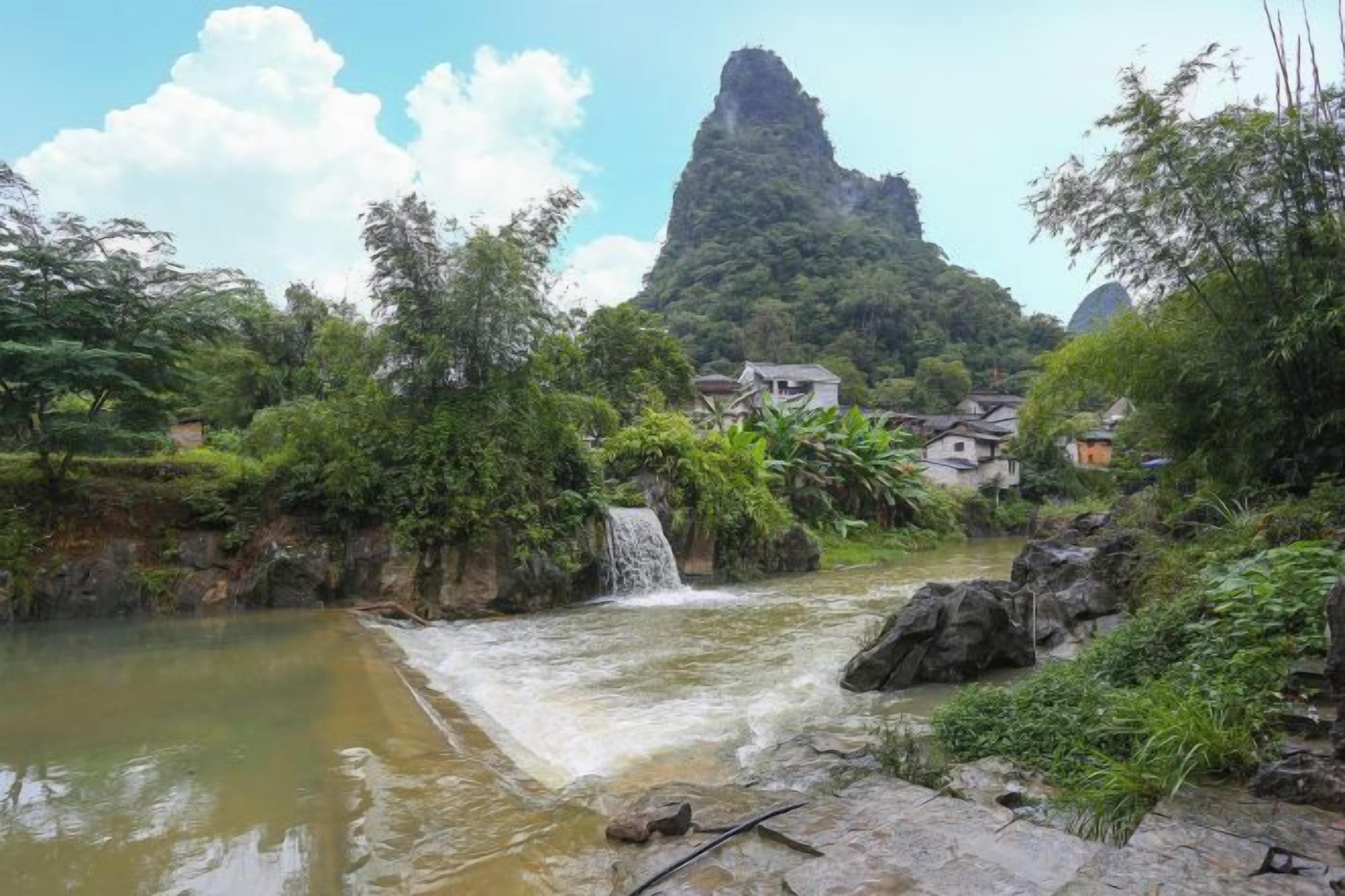 A river with a waterfall, surrounded by greenery and a mountain in the background.