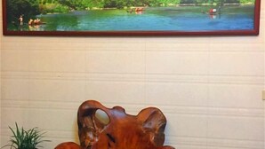 Un cuadro enmarco con una escena de río, barcos y gente, una escultura de madera, una planta en maceta y un bote en un estante.
