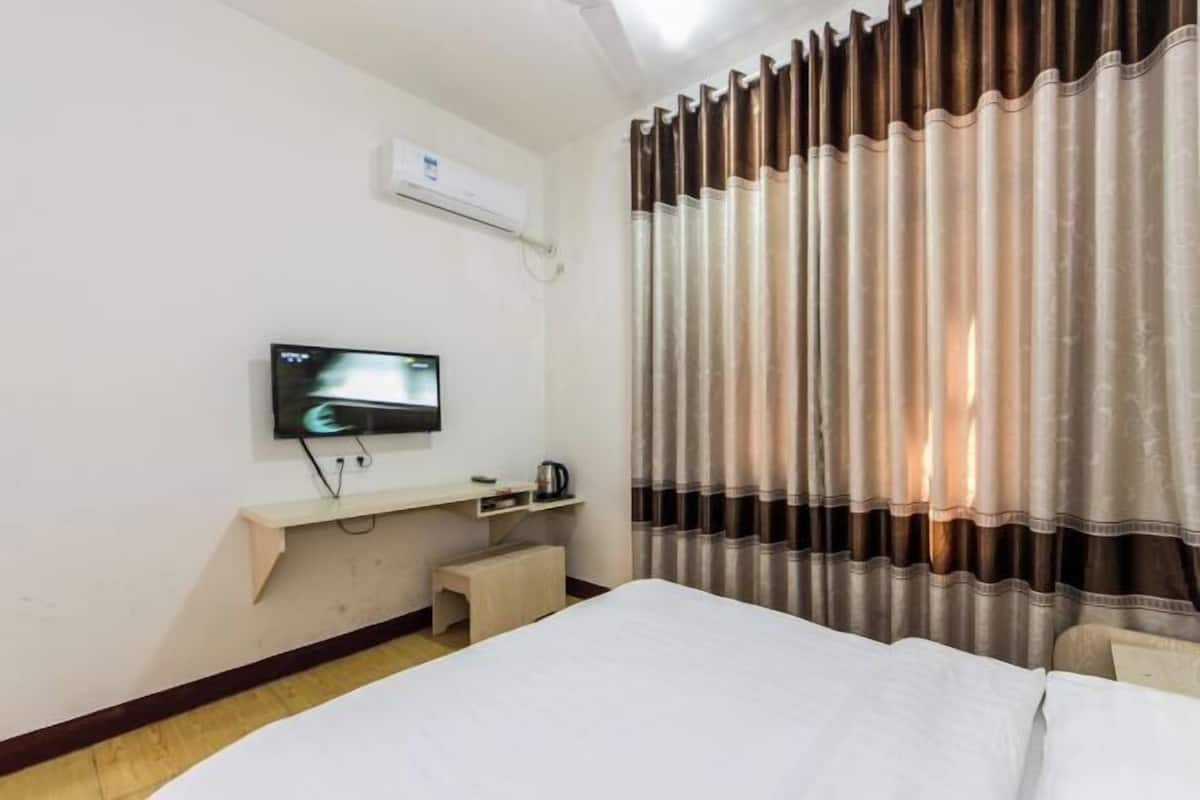 A hotel room with a bed, a TV mounted on the wall, a small table with a kettle, and a window with curtains.