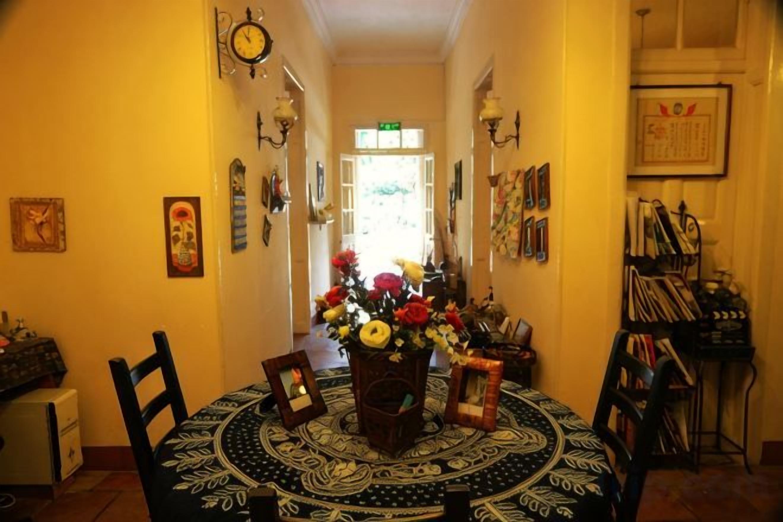 Uma área de refeições com uma mesa redonda, cadeiras e um vaso de flores. Há relógios fixados na parede e quadros emoldurados.