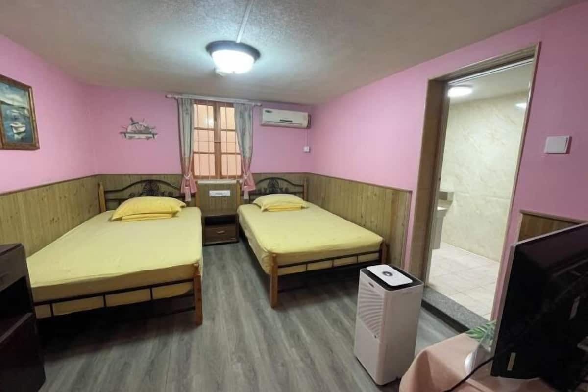 A room with two single beds, a desk with a computer, an air conditioner, and a window with curtains.