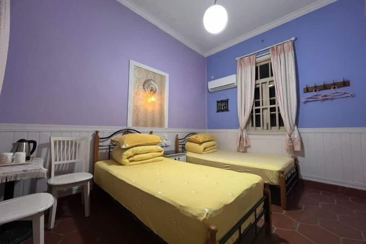 A room with two beds, a table with chairs, a hanging light fixture, and a window with curtains.