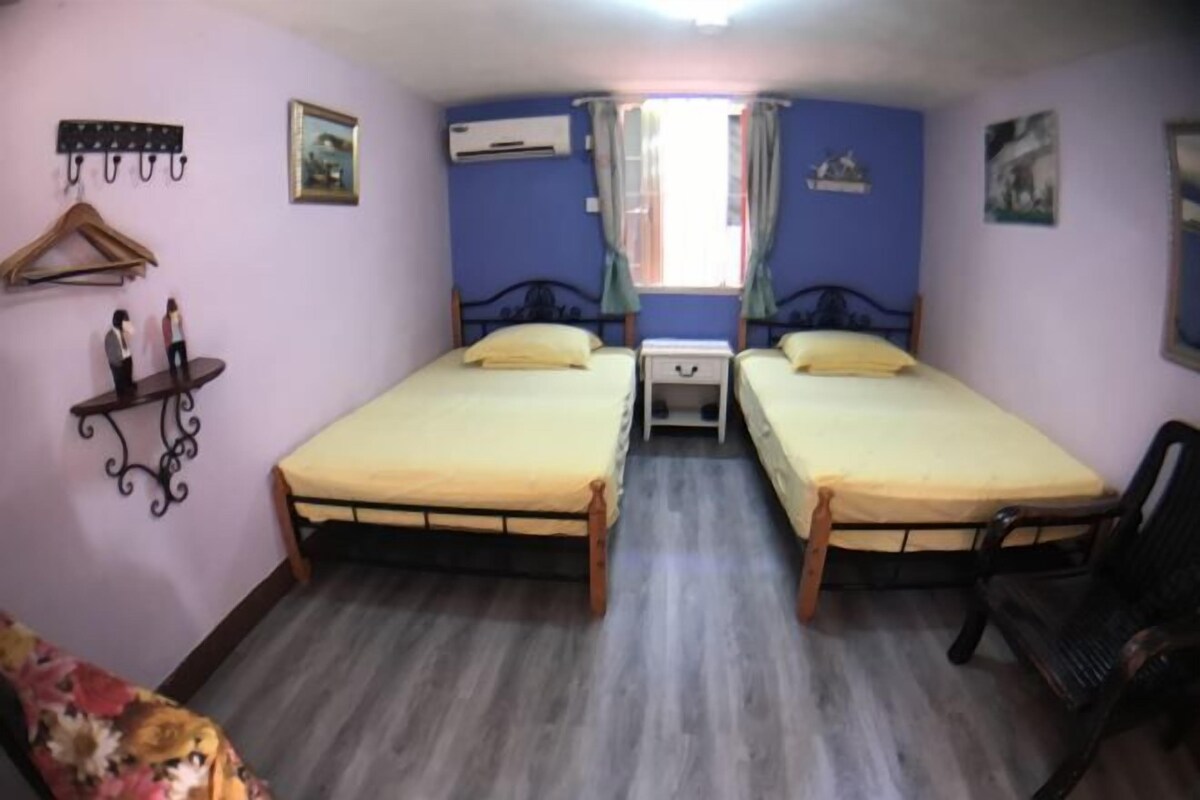 Una habitación con dos camas, piso de madera, una silla, perchero y ventana con cortinas.