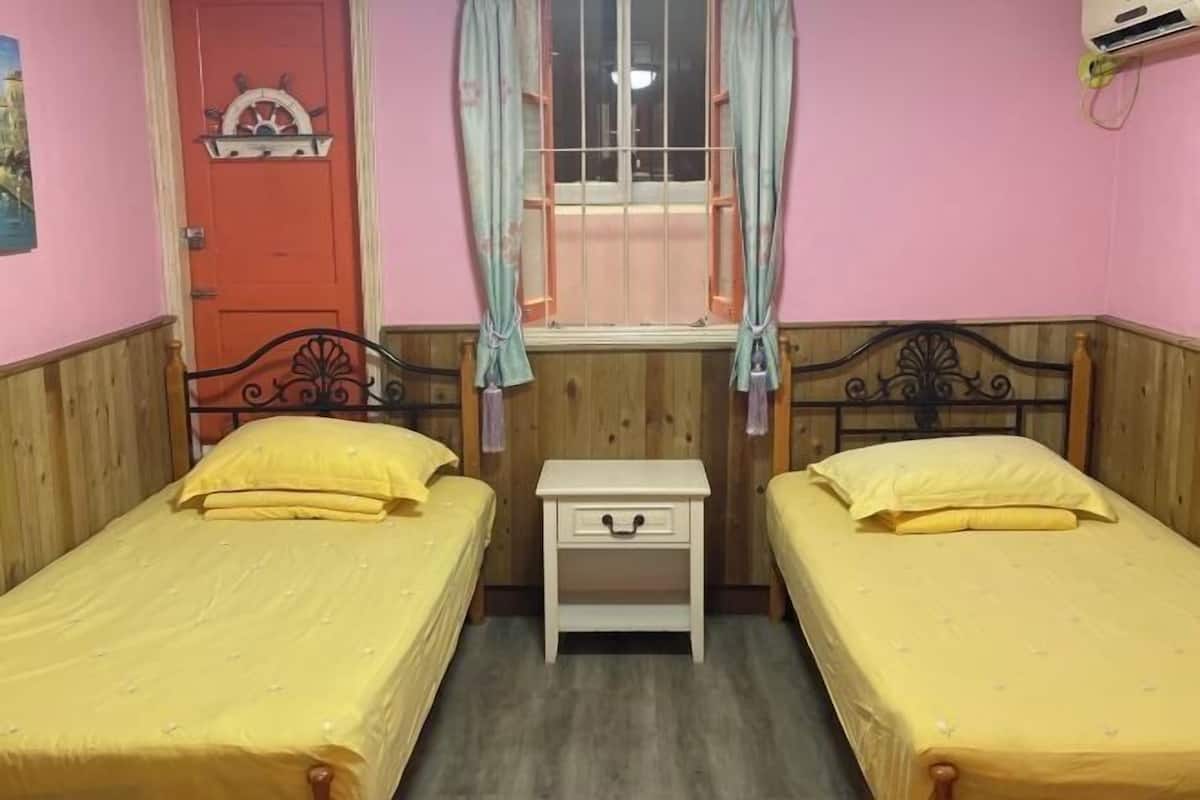 A room with two beds, a small white nightstand, a window with curtains, and a door with a decorative handle.