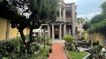A courtyard with a central pathway leading to a building with a balcony. There are potted plants and a small fountain.
