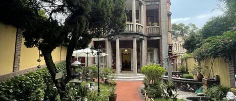 A courtyard with a central pathway leading to a building with a balcony. There are potted plants and a small fountain.