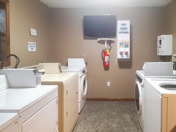 A laundry room with washing machines, dryers, a fire extinguisher, a television, and a soap and bleach softener dispenser.