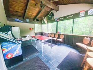 A room with a ping pong table, chairs, a refrigerator, and a window with a view of trees.