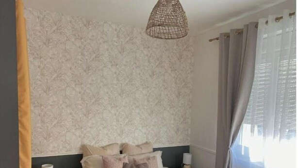 A bedroom with a bed, pillows, a bedside lamp, a hanging light fixture, and sheer curtains. - Le Quai des Marins (Châteauroux)