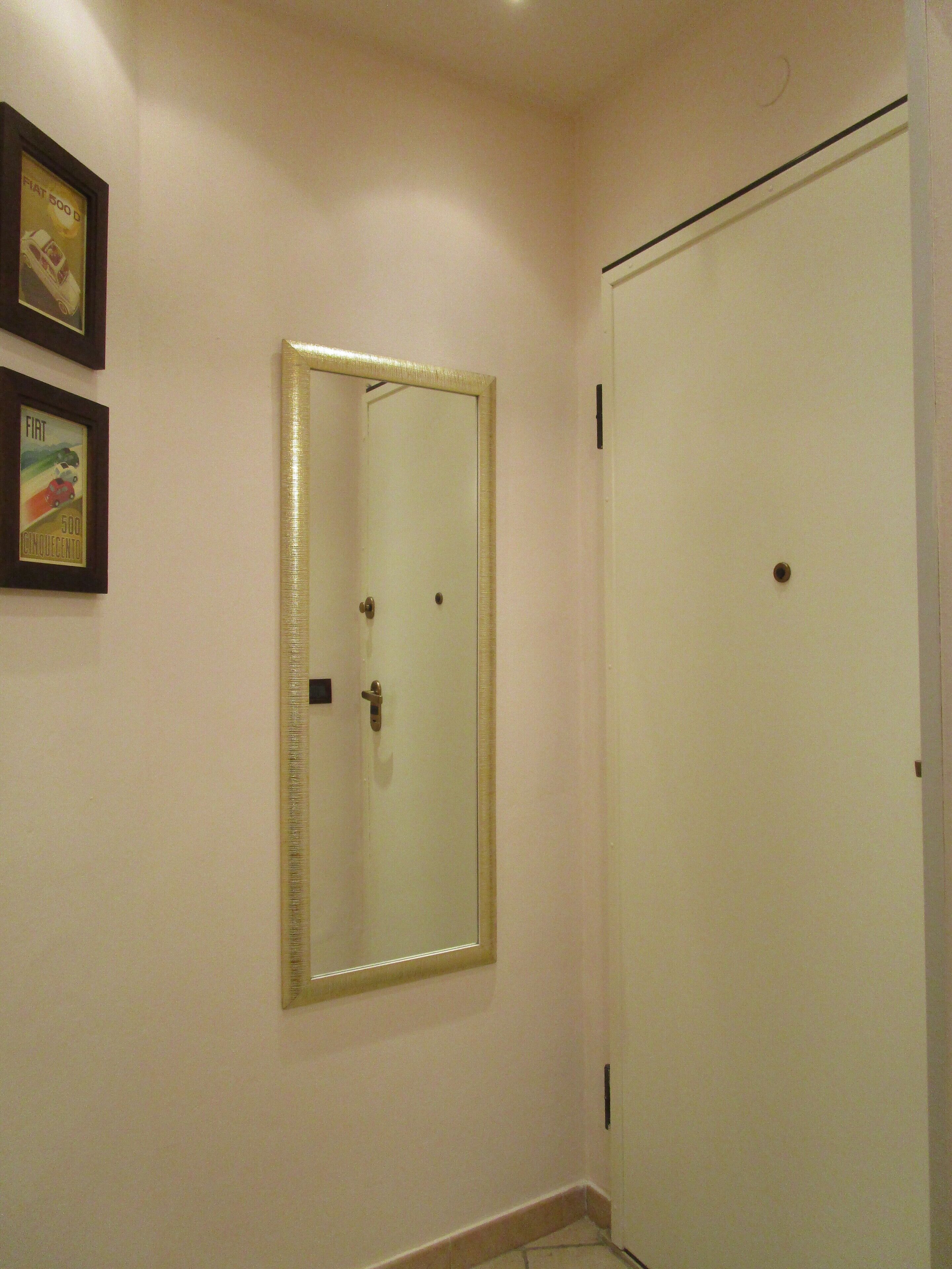 Uma porta branca com um espelho dourado acima, um pequeno quadro na parede à esquerda e uma parede branca lisa.