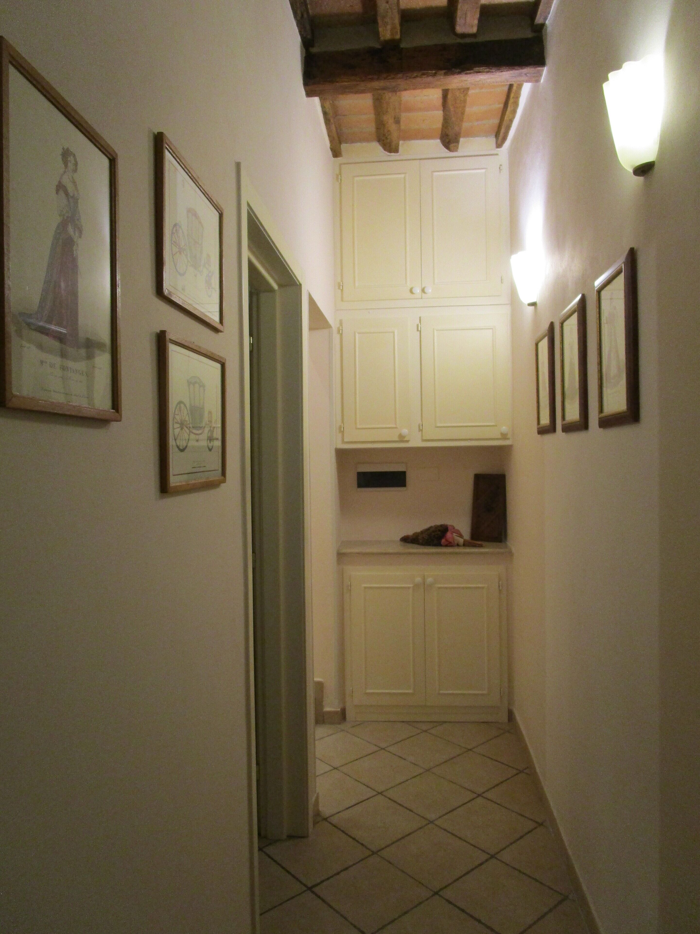 A narrow hallway with white tiled flooring, wooden beams on the ceiling, and several framed pictures on the walls.