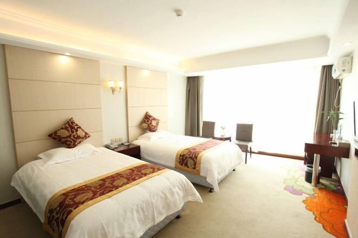 Une chambre d’hôtel avec deux lits, un bureau, une chaise, une grande fenêtre et un climatiseur fixé au mur.