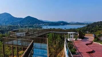 A wooden walkway with glass panels leading to a red-roofed area, overlooking a lake and distant mountains.