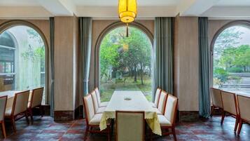 A dining area with a long table and chairs, large windows with curtains, and a hanging light fixture.