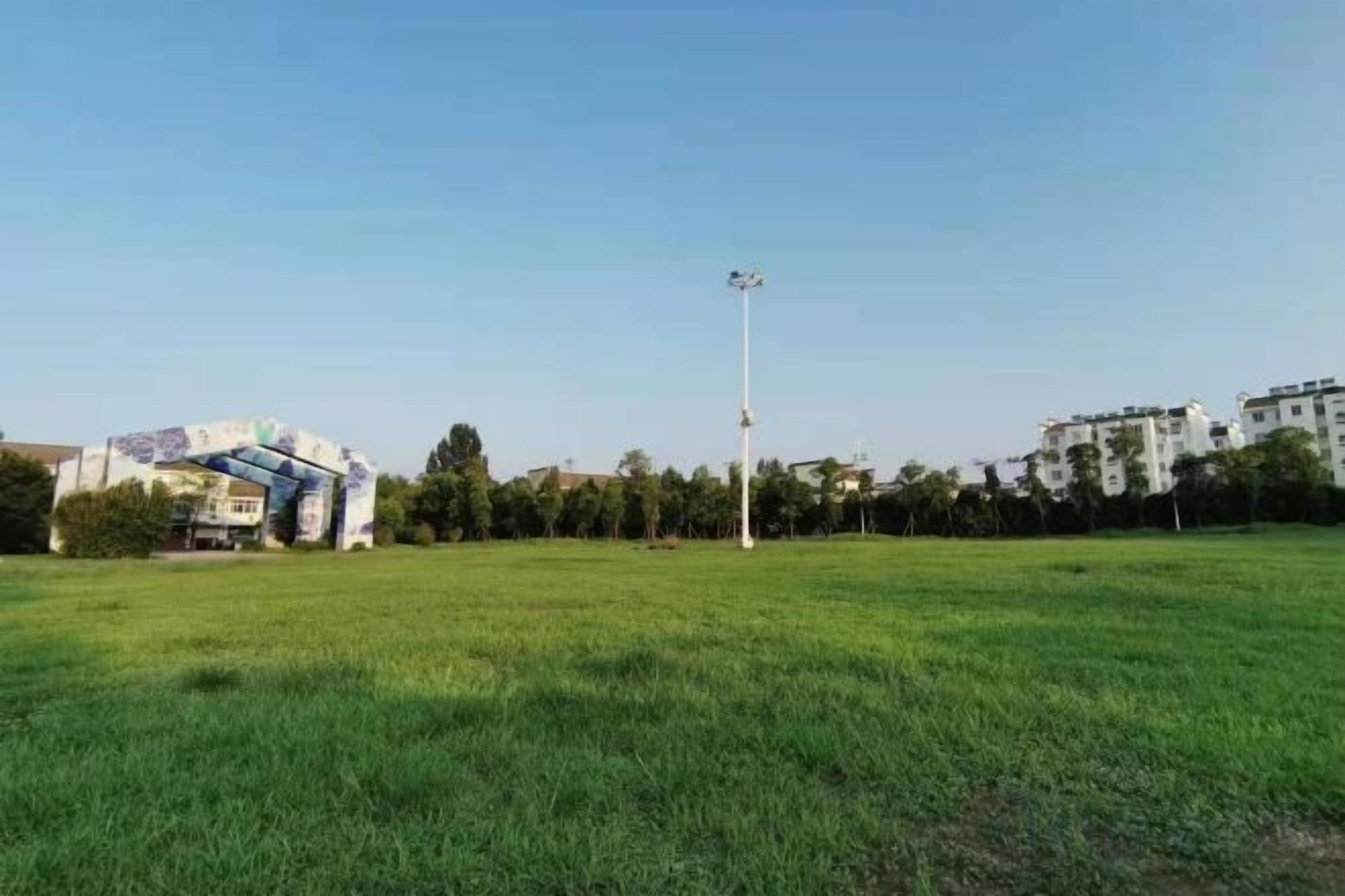 Un champ d’herbe avec un grand poteau d’éclairage, des bâtiments en arrière-plan et une structure colorée sur la gauche.